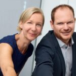 Zwei Personen lächeln in die Kamera, Bürohintergrund.