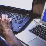 Person tippt auf Laptop-Tastatur vor zwei Bildschirmen.