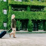 Woman walking with suitcase near ivy-covered building.