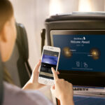 Passagier nutzt Smartphone neben Flugzeugschirm mit Reisedetails.