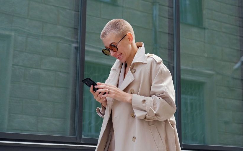 Eine gut gekleidete Frau mit einer getönten Brille überprüft ihr Telefon, während sie die Straße entlanggeht.