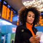 Mujer ejecutiva joven consultando el celular en un aeropuerto, rodeada por pantallas con información de vuelos.