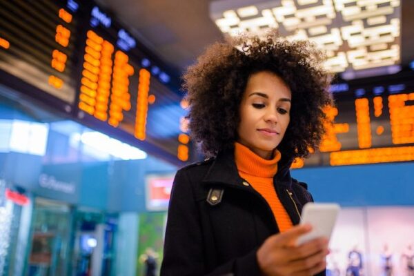 Mujer ejecutiva joven consultando el celular en un aeropuerto, rodeada por pantallas con información de vuelos.