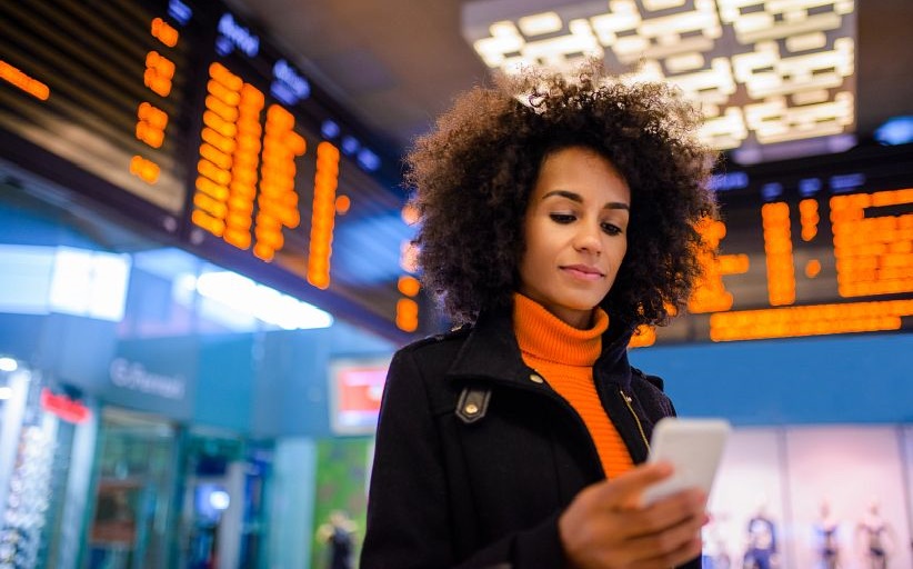 Mujer ejecutiva joven consultando el celular en un aeropuerto, rodeada por pantallas con información de vuelos.