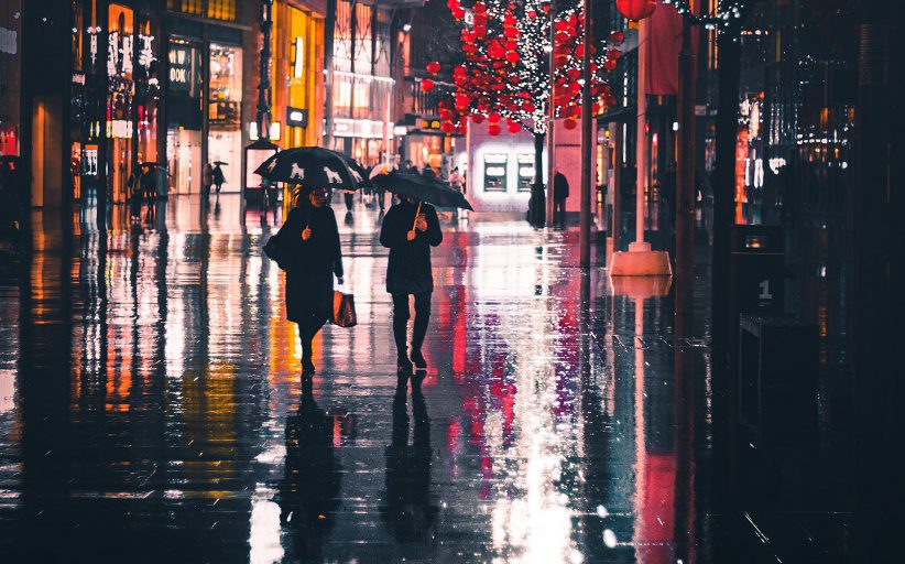 Hombre y mujer, elegantemente vestidos, caminan bajo la lluvia en una calle de la ciudad durante la noche. Ambos usan sombrilla.