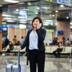 Mujer de negocios habla por teléfono celular mientras arrastra su maleta de cabina con ruedas por el suelo de un aeropuerto.