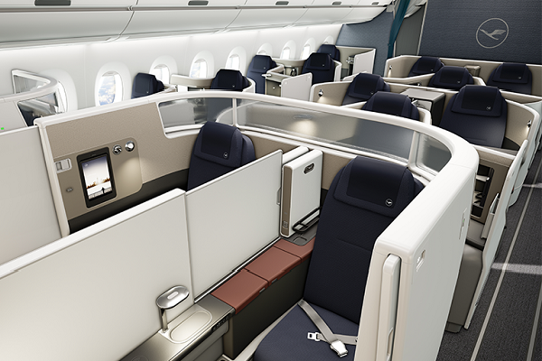Se aprecian varios asientos de avión, separados en cubículos, en una cabina de primera clase en cuya pared trasera se aprecia el logo de Lufthansa.