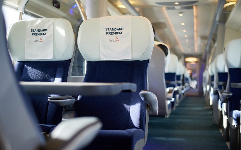 Interior de un vagón de tren de pasajeros. En los reposacabezas de dos de las sillas se lee Standard Premium y se ve el logo de la empresa Avanti West Coast.