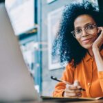 Mujer ejecutiva, con anteojos y blusa naranjada, mira a la cámara mientras sonríe, sentada frente a una laptop.