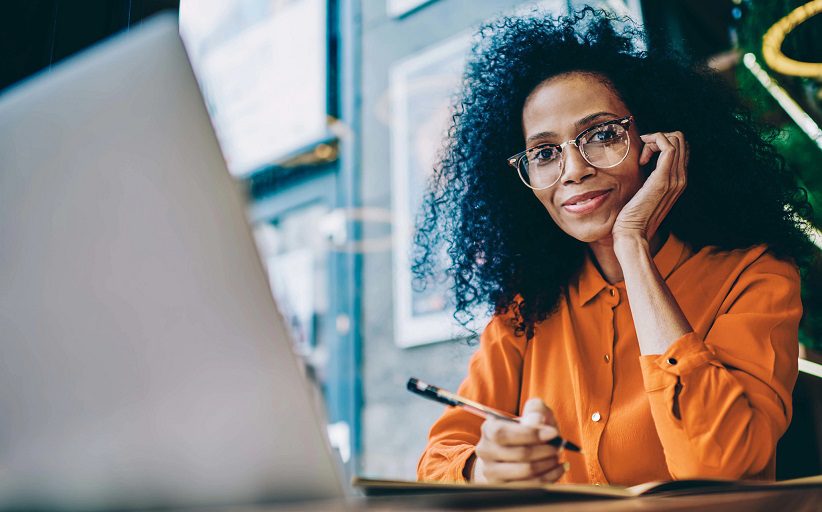 Mujer ejecutiva, con anteojos y blusa naranjada, mira a la cámara mientras sonríe, sentada frente a una laptop.