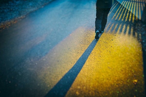 Se ven las piernas de alguien que camina, con yines y tenis. Una luz, por fuera del encuadre, proyecta en la calle la sombra de una reja y de sus piernas.