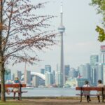 En primer plano, está el césped de un parque público con dos bancas y dos personas sentadas en cada banca. Al fondo, al otro lado del agua, se ve Toronto con la torre CN.