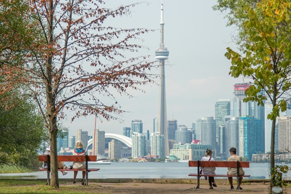 En primer plano, está el césped de un parque público con dos bancas y dos personas sentadas en cada banca. Al fondo, al otro lado del agua, se ve Toronto con la torre CN.