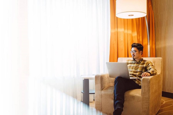 Un hombre trabaja en una laptop en el lobby de un hotel. Está sentado en un sillón bajo una enorme lámpara de pie.