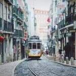Un tranvía avanza por el medio de una calle colonial en una ciudad portuguesa. A cada lado, se ven casas de dos plantas y, en el fondo, un edificio.