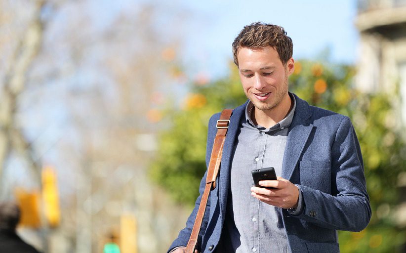 Hombre joven sonríe mientras consulta su celular al caminar por la calle.