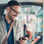 Un hombre de negocios sonríe mientras revisa la pantalla de su celular y permanece de pie en el vagón de un tren subterráneo.