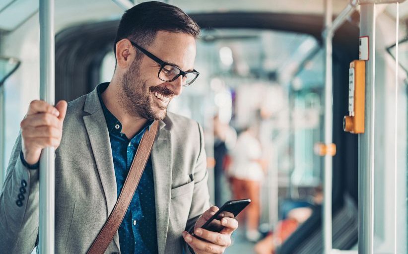 Un hombre de negocios sonríe mientras revisa la pantalla de su celular y permanece de pie en el vagón de un tren subterráneo.