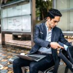 Hombre de negocios extrae un documento de su mochila mientras trabaja sentado en un espacio púbico al exterior, con una laptop en sus rodillas.