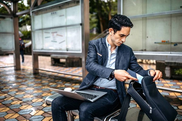 Hombre de negocios extrae un documento de su mochila mientras trabaja sentado en un espacio púbico al exterior, con una laptop en sus rodillas.