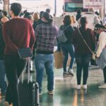 Varios pasajeros, algunos de ellos con maleta viajera o morral, aguardan pacientemente de pie en una sala de espera.