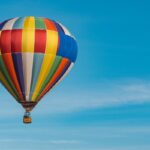 Un globo de múltiples colores vuela en un cielo otoñal, limpio y azul. En la canastilla viajan varias personas.