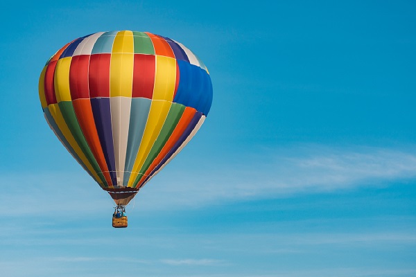 Un globo de múltiples colores vuela en un cielo otoñal, limpio y azul. En la canastilla viajan varias personas.
