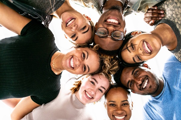Un grupo de siete personas, entre mujeres y hombres de diferentes razas, forman un círculo juntando sus rostros y sonríen mientras miran a la cámara, ubicada debajo de ellos.