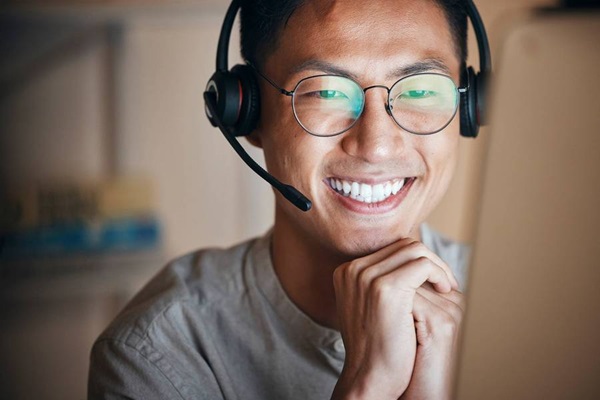 Joven con anteojos mira la pantalla de un computador mientras sonríe. Usa una diadema con audífonos y un micrófono incorporado.