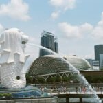 Una figura blanca de piedra, con cabeza de león, y torso y cola de sirena, vierte agua en una fuente. Al fondo, se ven diversos edificios de la ciudad-estado de Singapur.
