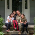 Un soldado vistiendo el uniforme del Ejército de Estados Unidos abraza a su esposa y sus dos hijos sentados en el porche de su casa.