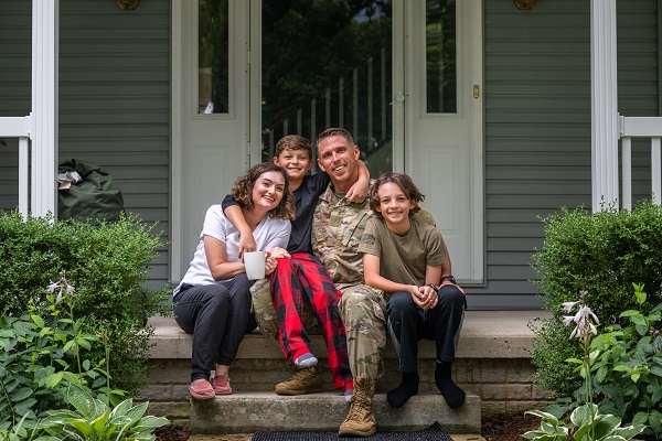 Un soldado vistiendo el uniforme del Ejército de Estados Unidos abraza a su esposa y sus dos hijos sentados en el porche de su casa.