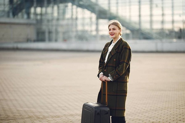 Una mujer de negocios sonríe mientras sostiene con ambas manos, sosteniéndola de la manija extendida, su maleta de cabina, que se encuentra en el suelo.