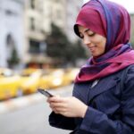 Joven mujer en la calle usando un hiyab violeta y consultando el celular mientras sonríe.