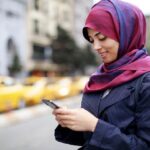 Joven mujer en la calle usando un hiyab violeta y consultando el celular mientras sonríe.