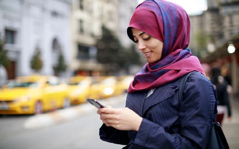 Joven mujer en la calle usando un hiyab violeta y consultando el celular mientras sonríe.