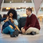 Una pareja de adultos está sentada en el suelo de lo que parece un aeropuerto. Tienen sus maletas detrás de ellos. Ella carga y abraza a una niña que parece estar enferma.