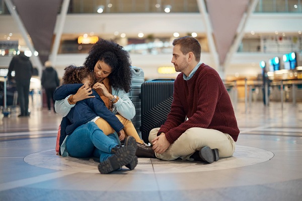 Una pareja de adultos está sentada en el suelo de lo que parece un aeropuerto. Tienen sus maletas detrás de ellos. Ella carga y abraza a una niña que parece estar enferma.