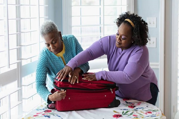 Dos mujeres tratan de cerrar juntas una maleta de cabina con ruedas, y de color rojo, que se encuentra sobre una mesa.