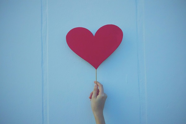 Una mano sostiene, contra un fondo blanco, un corazón rojo de papel pegado en un palillo de madera.