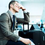 Hombre joven de negocios, sentado en la sala de espera de un aeropuerto, se lleva una mano al rostro en un gesto de preocupación