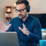 Hombre con gafas y audífonos explica algo mirando a la pantalla de una laptop.