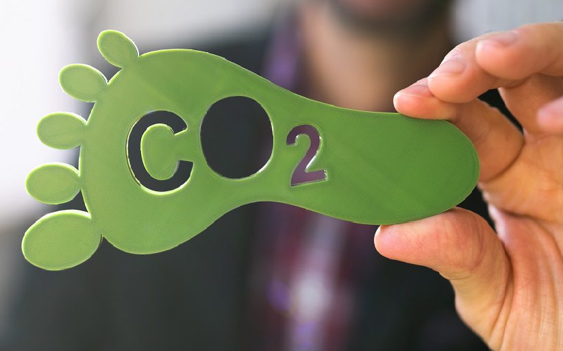 Una mano sostiene, en primer plano, un objeto de madera de color verde en forma de huella de pie humana. En este, puede leerse la inscripción CO2.
