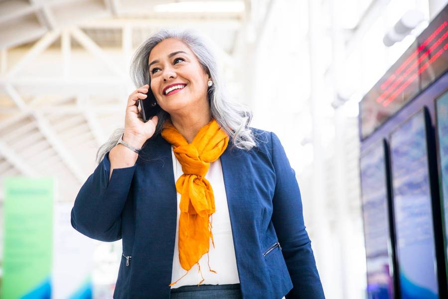 Mujer de negocios sonríe mientras habla por celular y camina por el corredor de lo que parece ser un aeropuerto.