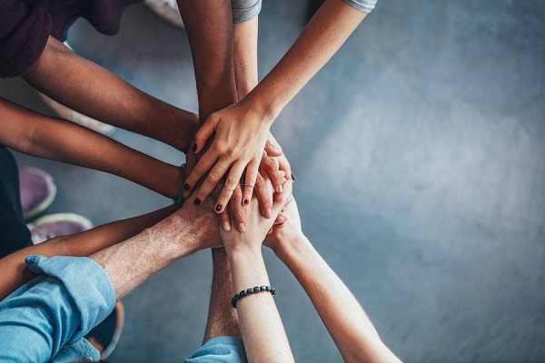 Se ven los brazos y manos de cinco hombres y mujeres que juntan sus manos en ademán de trabajo en equipo y celebración.