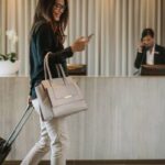 Mujer joven sonríe mientras mira el celular y arrastra una maleta de cabina en el lobby de un hotel. Al fondo, en el mostrador, una empleada del hotel responde el teléfono.