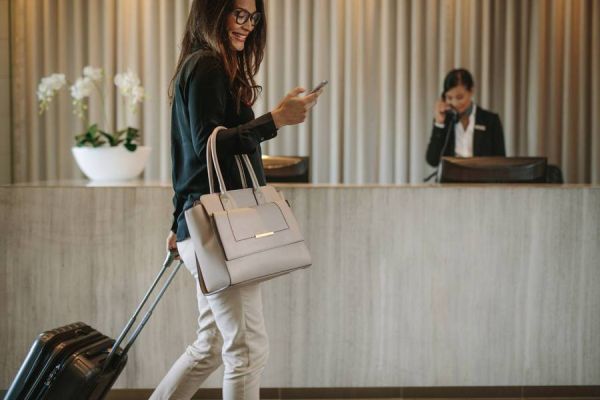 Mujer joven sonríe mientras mira el celular y arrastra una maleta de cabina en el lobby de un hotel. Al fondo, en el mostrador, una empleada del hotel responde el teléfono.