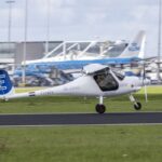 Un pequeño avión elécrico, de colores azul y blanco, despega de una pista de aeropuerto. Al fondo, puede verse un avión con el logo de KLM.