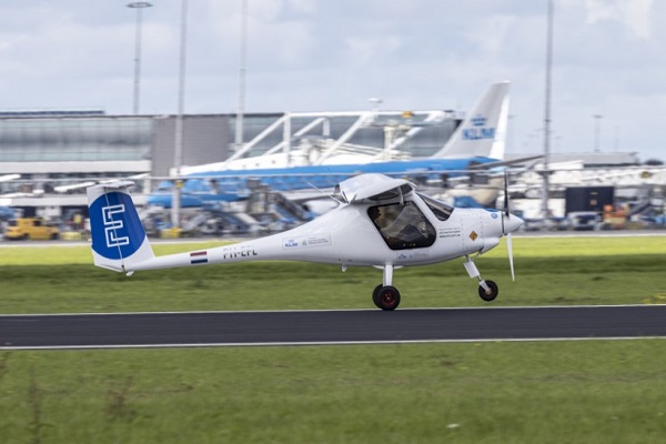 Un pequeño avión elécrico, de colores azul y blanco, despega de una pista de aeropuerto. Al fondo, puede verse un avión con el logo de KLM.