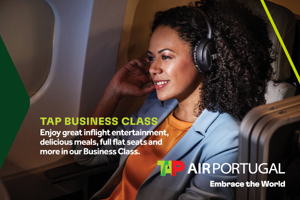 Una mujer viaja en un avión, sentada al lado de la ventanilla y escuchando a través de audífonos mientras sonríe.